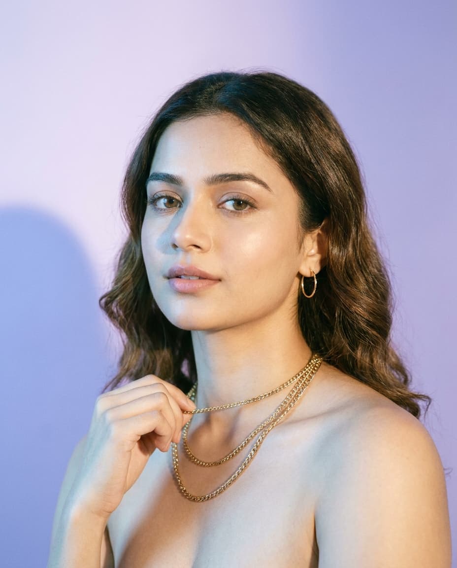 Woman adjusting layered gold necklaces against a soft lavender studio backdrop